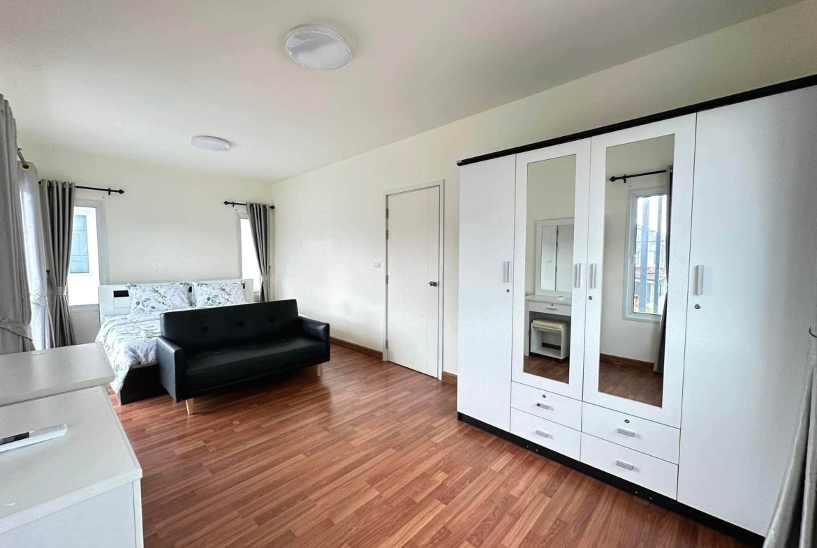 Bright contemporary bedroom with white built-in wardrobe featuring mirrored doors, a black sofa, and a bed against a white wall with wood flooring and windows with gray curtains.
