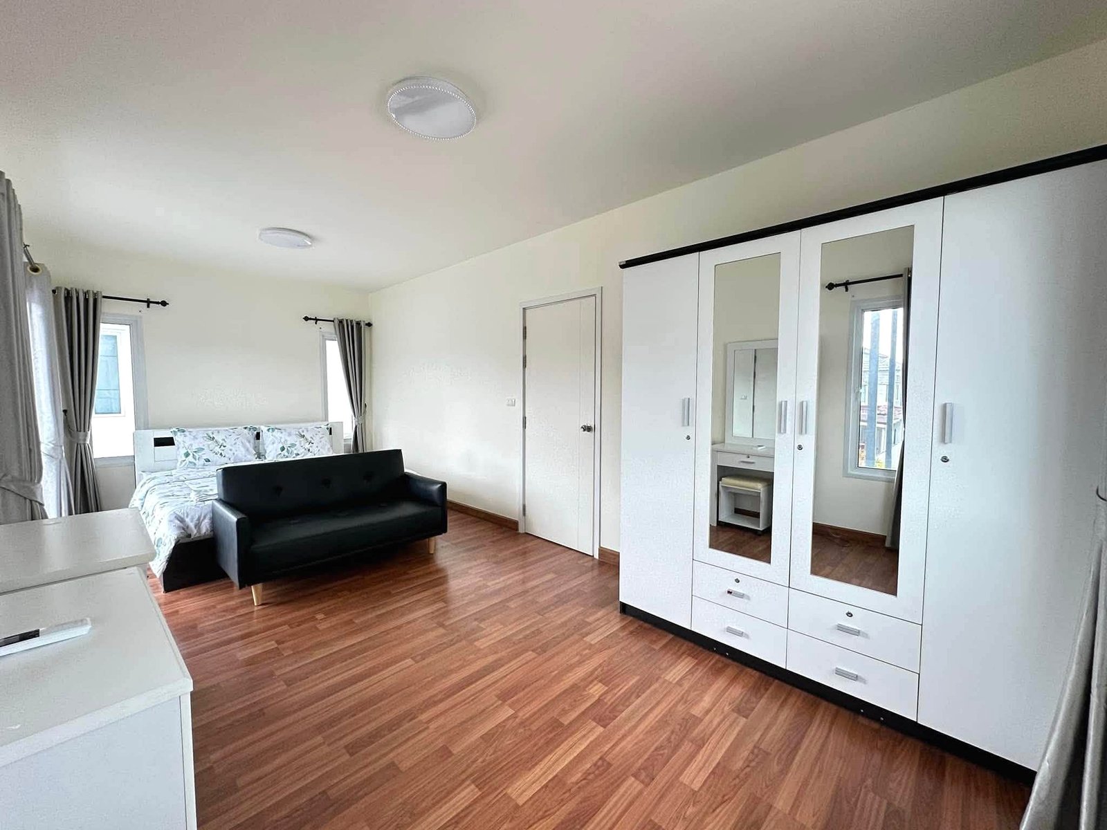 Pruksa_Nara_Pattaya_Rent_Rdl351_10 Bright contemporary bedroom with white built-in wardrobe featuring mirrored doors, a black sofa, and a bed against a white wall with wood flooring and windows with gray curtains.