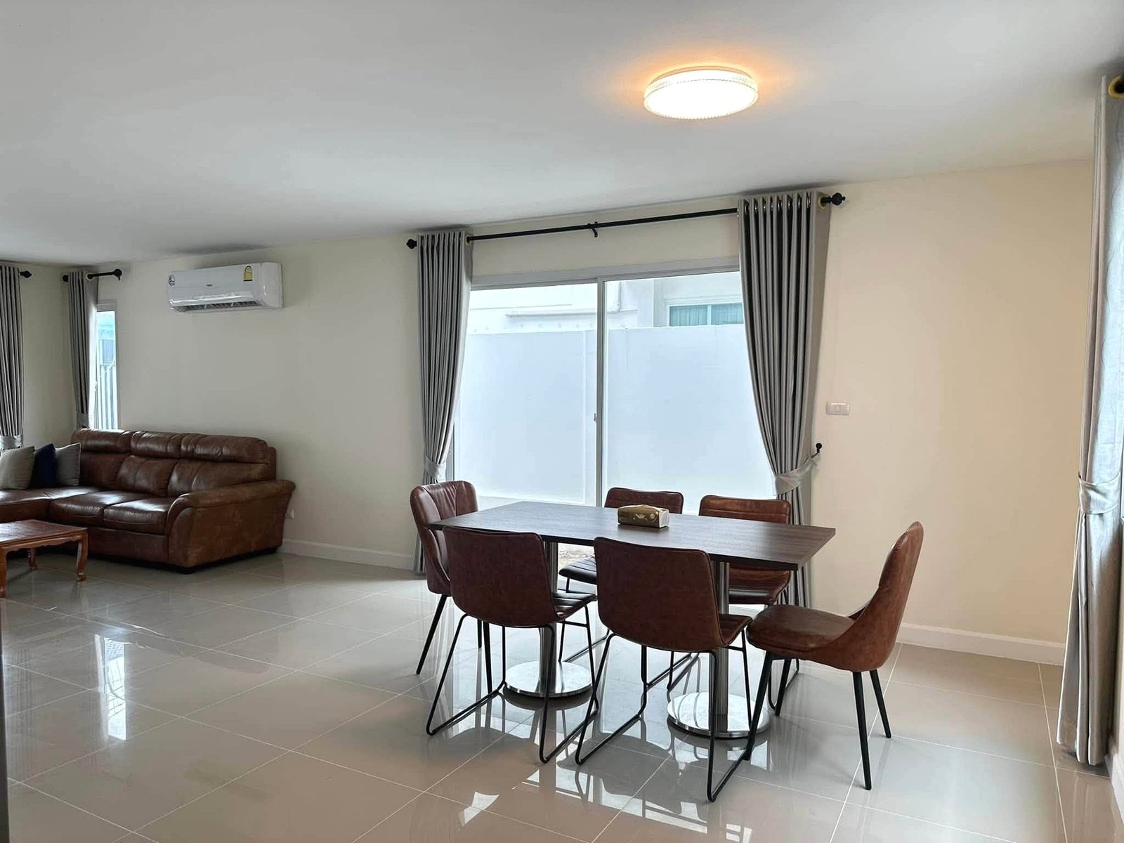 Pruksa_Nara_Pattaya_Rent_Rdl351_14 Open living and dining area featuring a brown leather sofa, a six-seat dining table with chairs, tiled floor, and large windows with gray curtains along the wall with an air conditioner above.