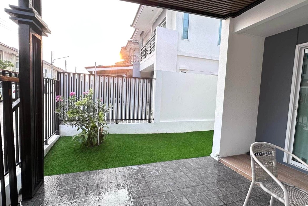 Patio area with artificial grass, a flowering shrub, black metal fence, and a sliding glass door to a modern house.