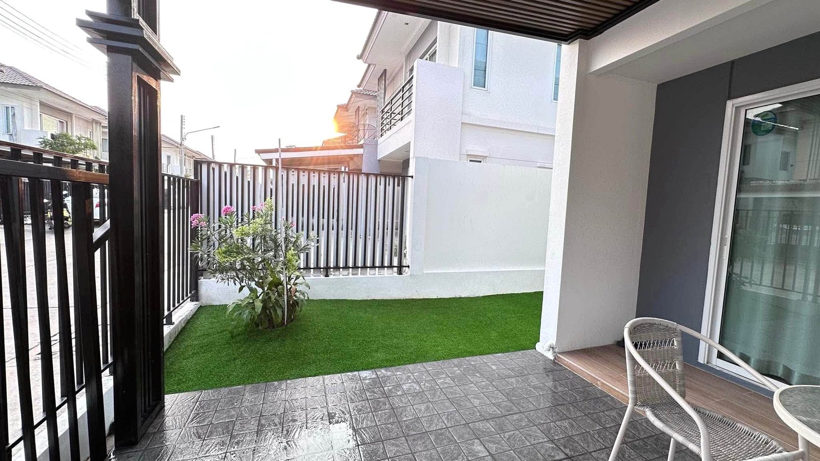 Pruksa_Nara_Pattaya_Rent_Rdl351_17 Patio area with artificial grass, a flowering shrub, black metal fence, and a sliding glass door to a modern house.