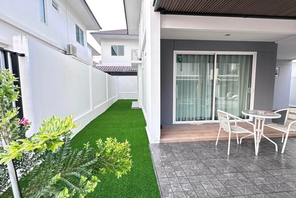 Narrow outdoor courtyard between white walls with a green artificial lawn, a sliding glass door, and a small white metal table with chairs on a tiled patio.