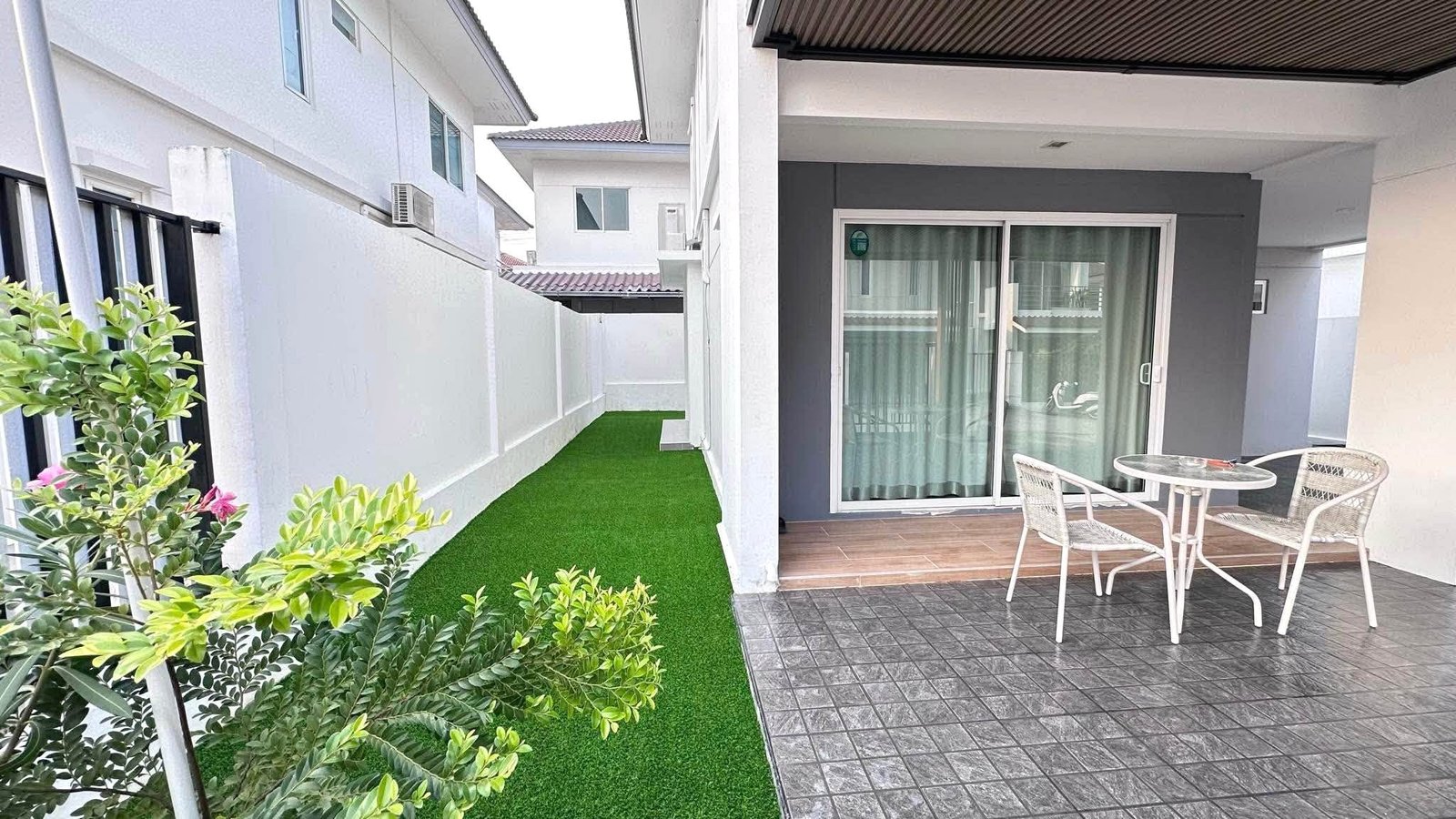 Pruksa_Nara_Pattaya_Rent_Rdl351_2 Narrow outdoor courtyard between white walls with a green artificial lawn, a sliding glass door, and a small white metal table with chairs on a tiled patio.