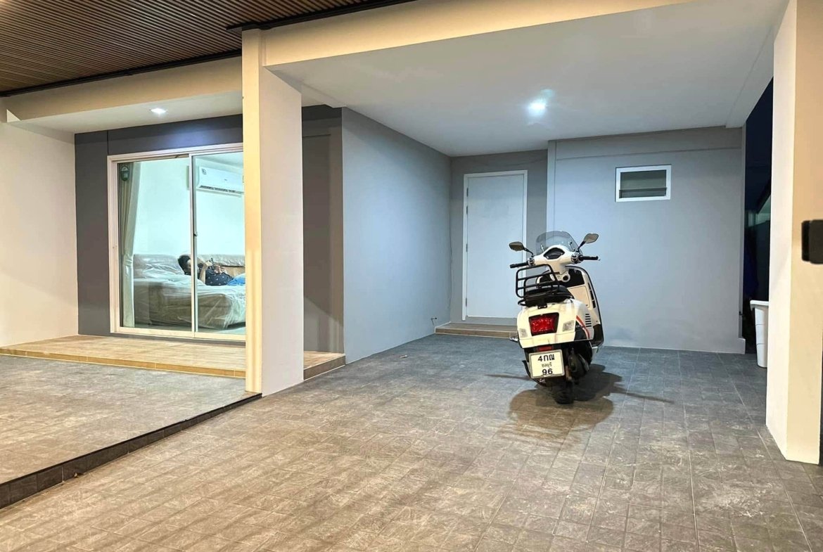 White scooter parked in a covered garage area beside a gray wall; sliding glass doors reveal a bedroom inside to the left.