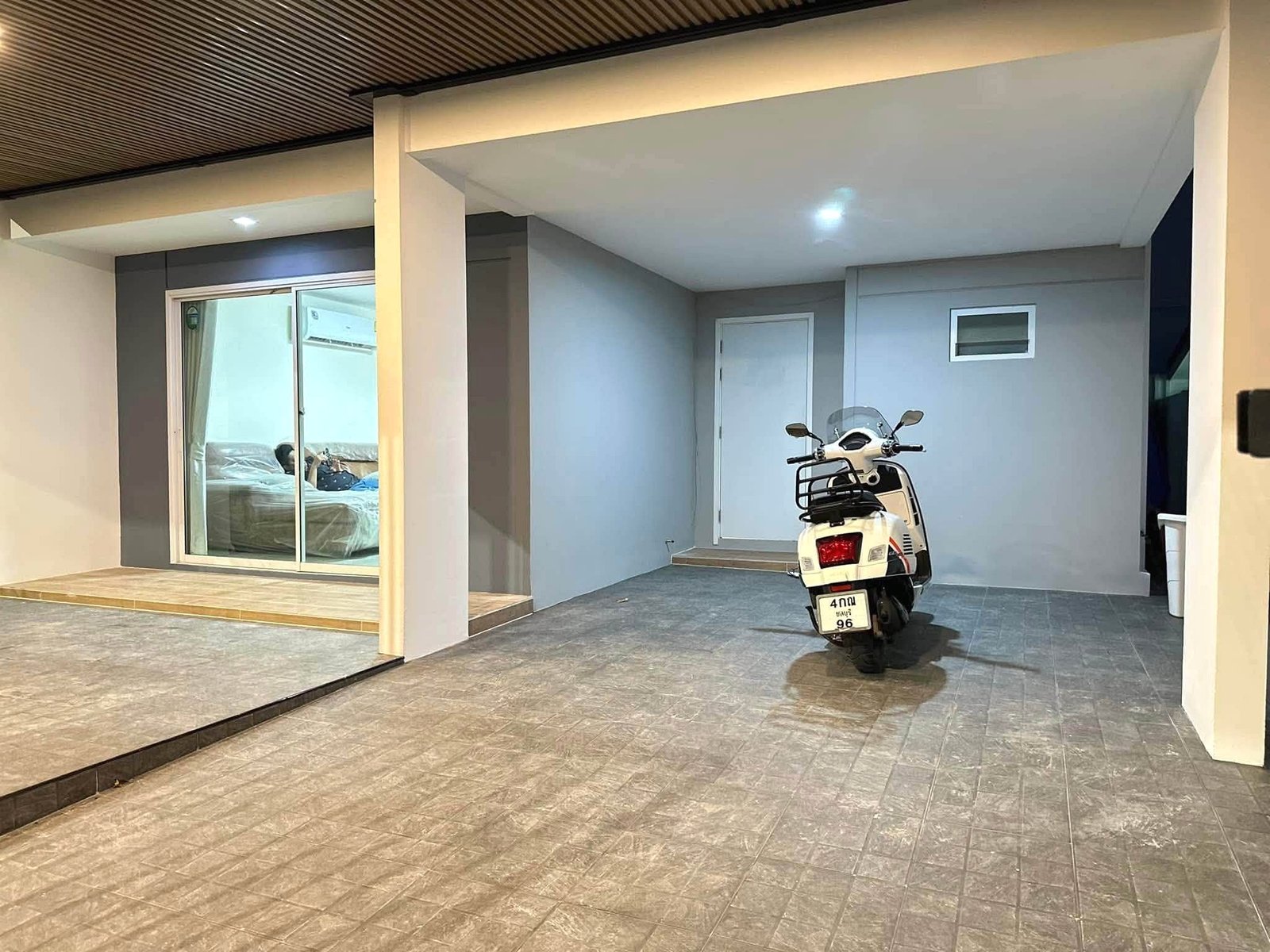 Pruksa_Nara_Pattaya_Rent_Rdl351_20 White scooter parked in a covered garage area beside a gray wall; sliding glass doors reveal a bedroom inside to the left.