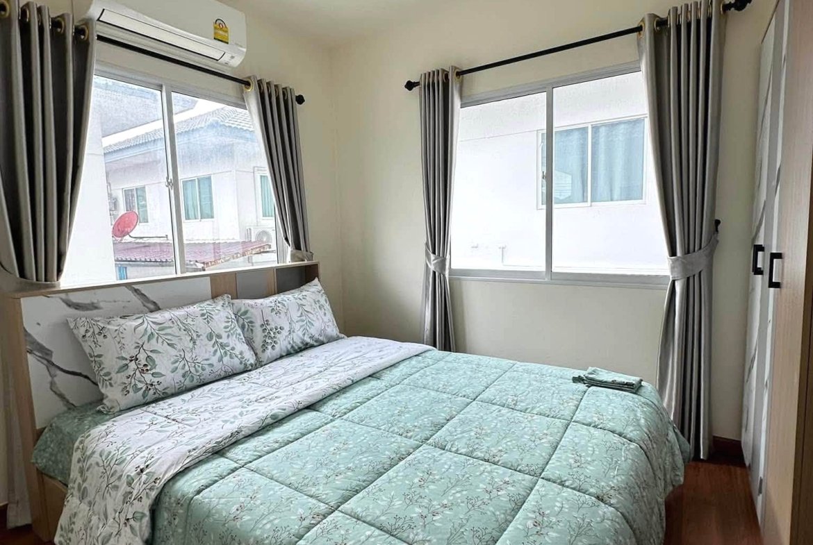 Small bedroom with a mint-green floral quilted bedspread, pillows, and wooden floor; two windows with gray curtains and a wall AC unit above the bed.
