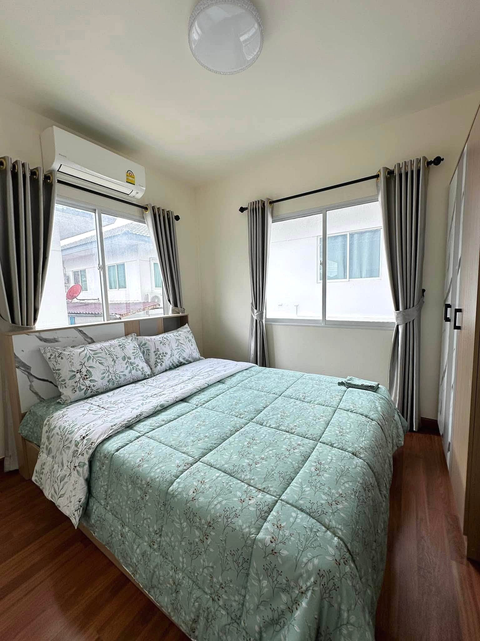 Pruksa_Nara_Pattaya_Rent_Rdl351_5 Small bedroom with a mint-green floral quilted bedspread, pillows, and wooden floor; two windows with gray curtains and a wall AC unit above the bed.