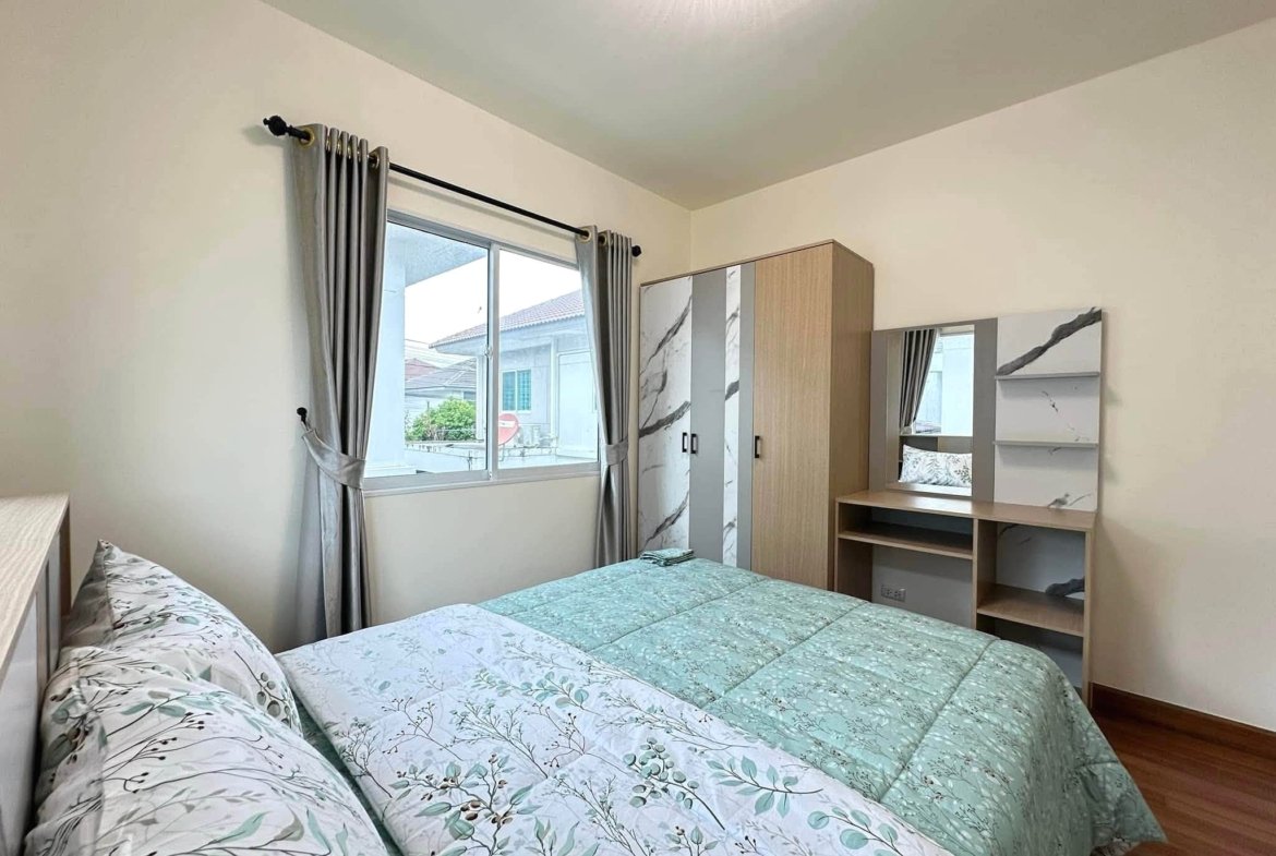 Two twin beds with green botanical-pattern quilts in a bright bedroom with a window and gray curtains on the left side of the room.