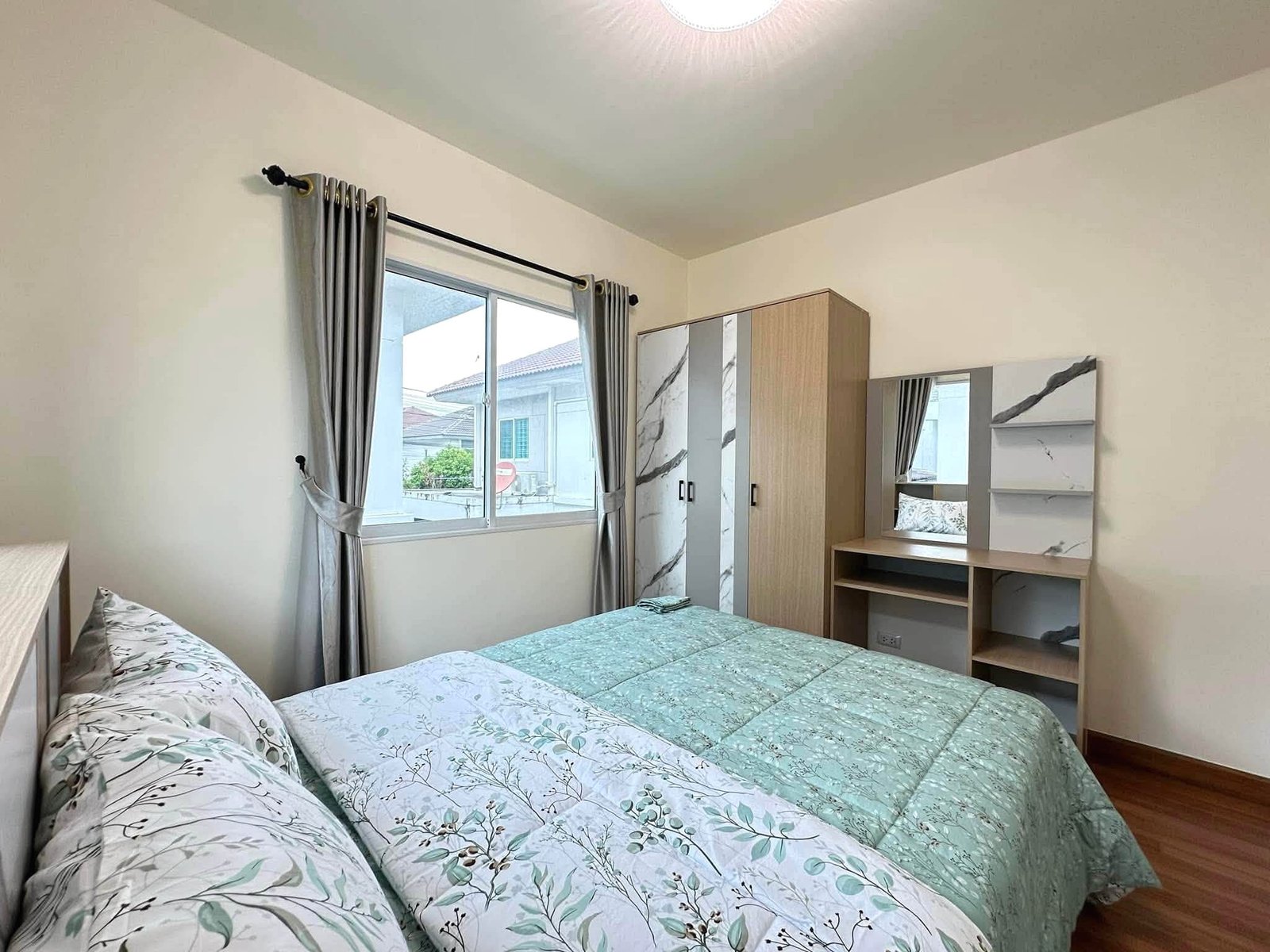 Pruksa_Nara_Pattaya_Rent_Rdl351_6 Two twin beds with green botanical-pattern quilts in a bright bedroom with a window and gray curtains on the left side of the room.