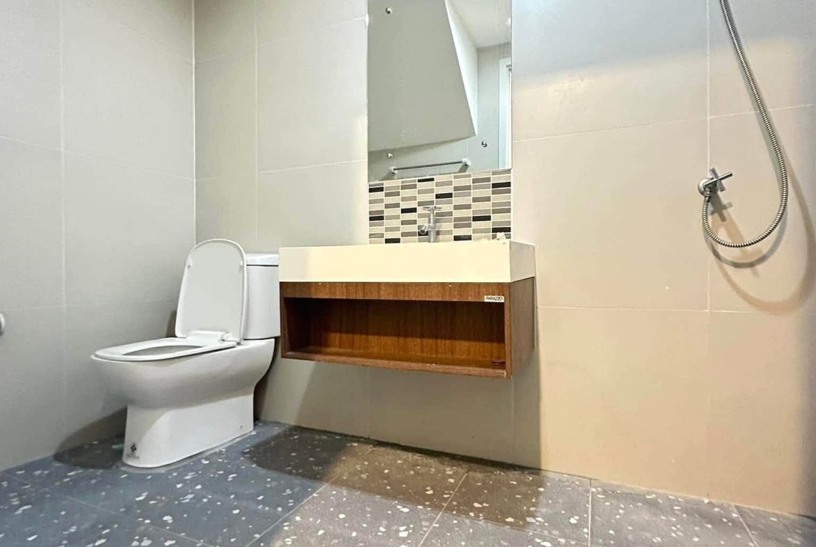 Modern bathroom with a white toilet, a wooden floating vanity, and a mirror framed by a mosaic tile band; handheld shower on the right edge.