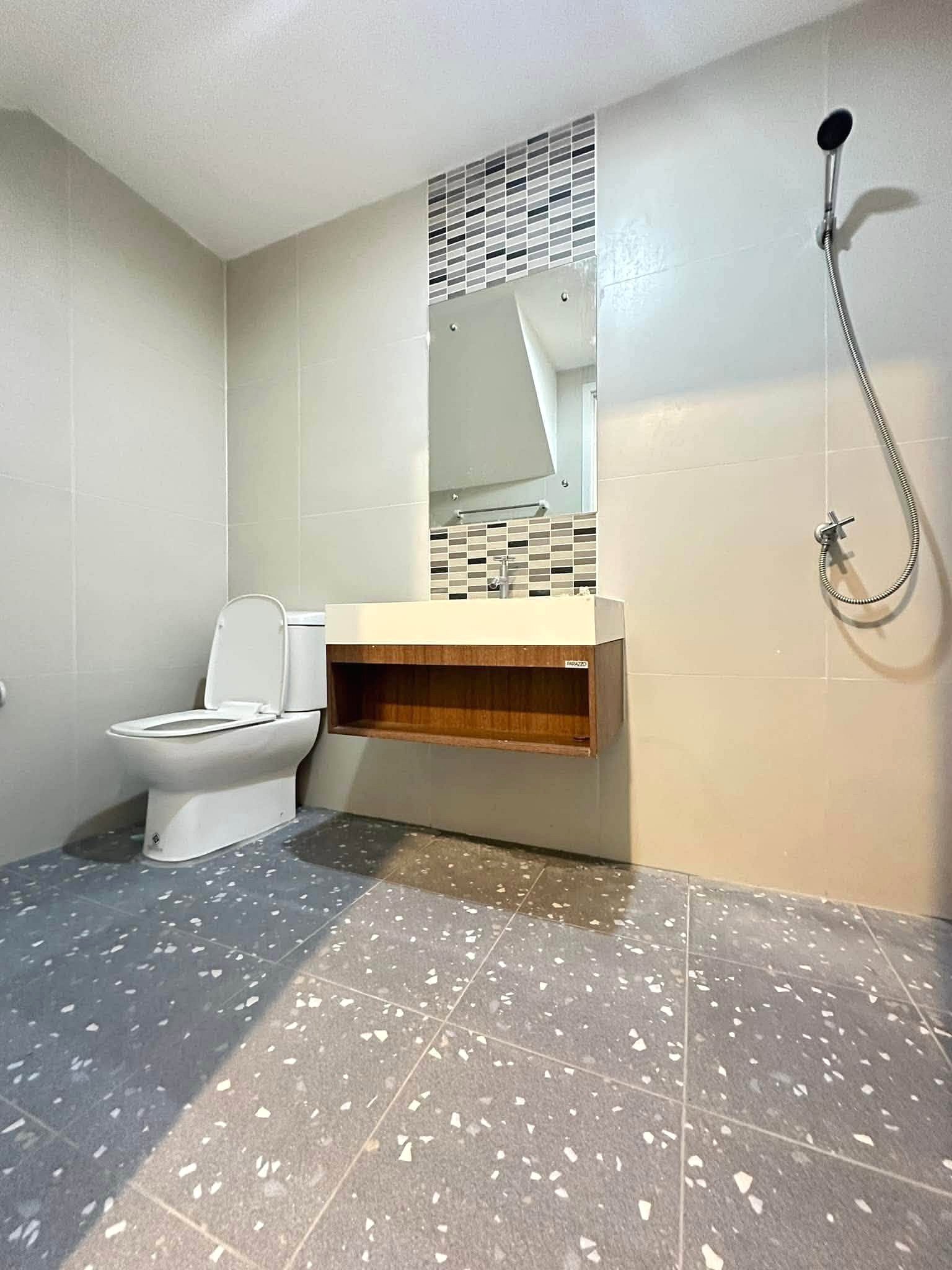 Pruksa_Nara_Pattaya_Rent_Rdl351_7 Modern bathroom with a white toilet, a wooden floating vanity, and a mirror framed by a mosaic tile band; handheld shower on the right edge.