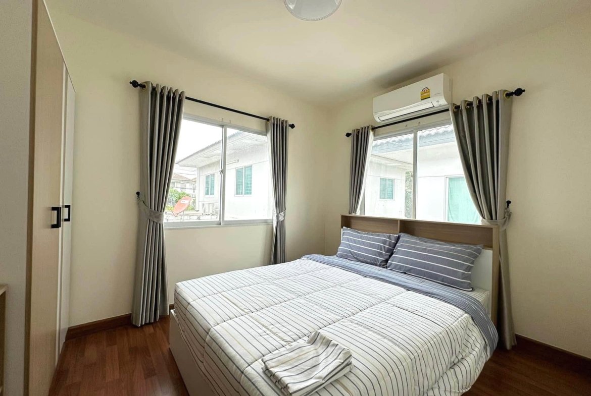 Two single beds with striped bedding placed side by side in a small bedroom, featuring gray curtains, wooden floor, and a wall-mounted air conditioner above the window.