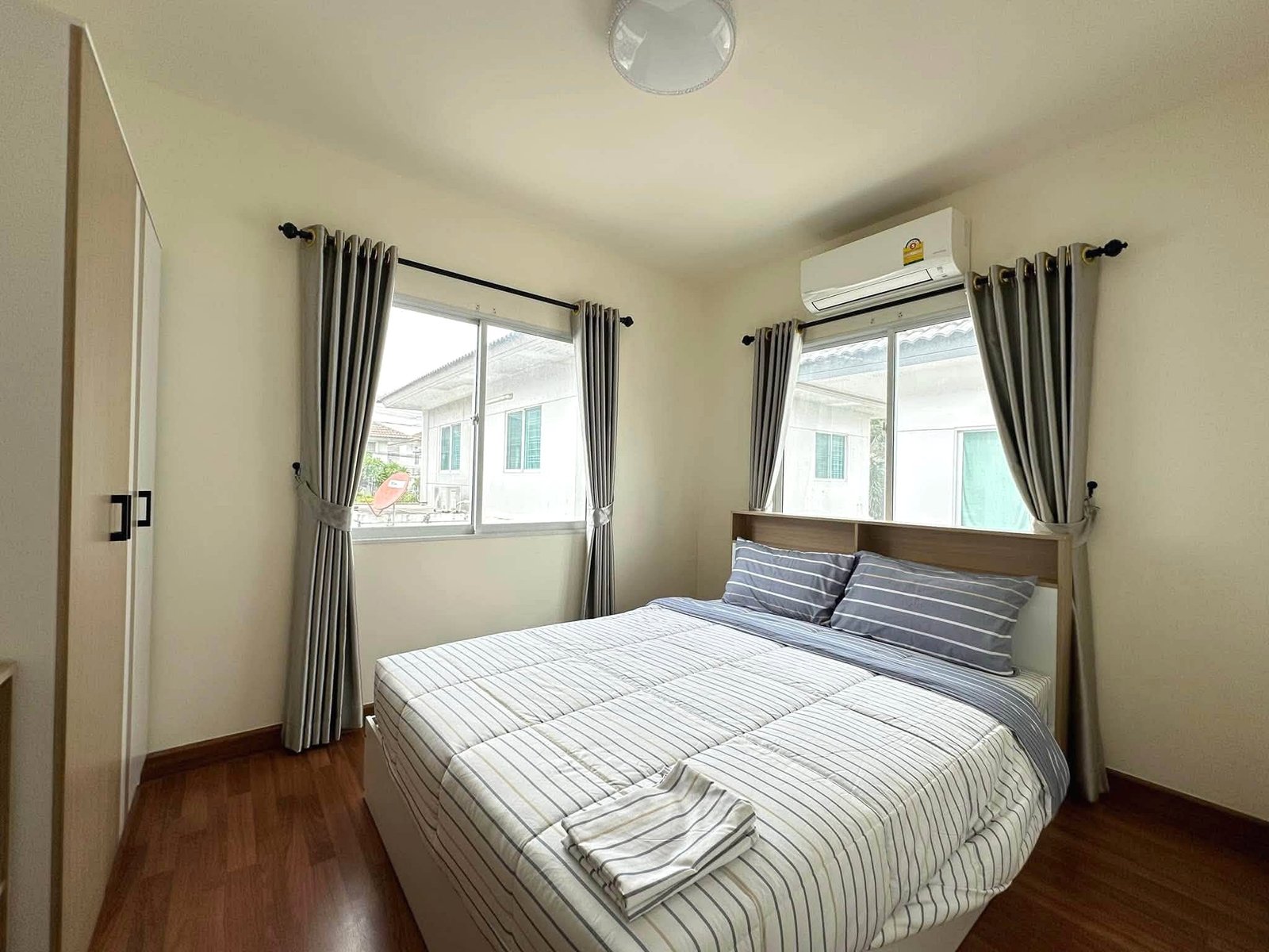 Pruksa_Nara_Pattaya_Rent_Rdl351_8 Two single beds with striped bedding placed side by side in a small bedroom, featuring gray curtains, wooden floor, and a wall-mounted air conditioner above the window.