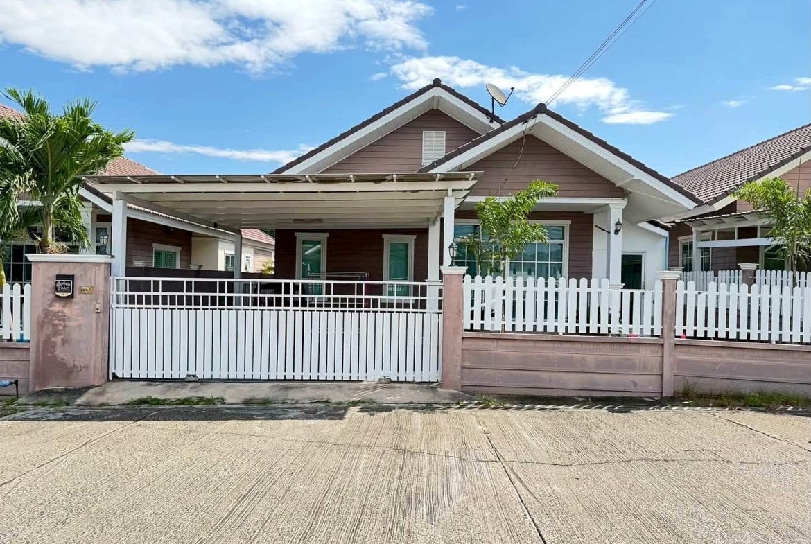 Front view of a single-story house with a white picket fence, pinkish-brown siding, and a driveway gate under a bright blue sky with clouds, a satellite dish on the roof.
