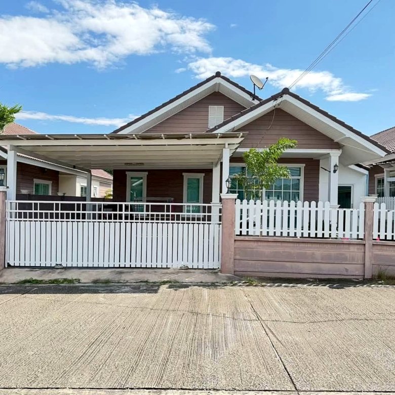 Front view of a single story house with a white picket fence pinkish brown siding and a driveway gate under a bright blue sky with clouds a satellite dish on the roof