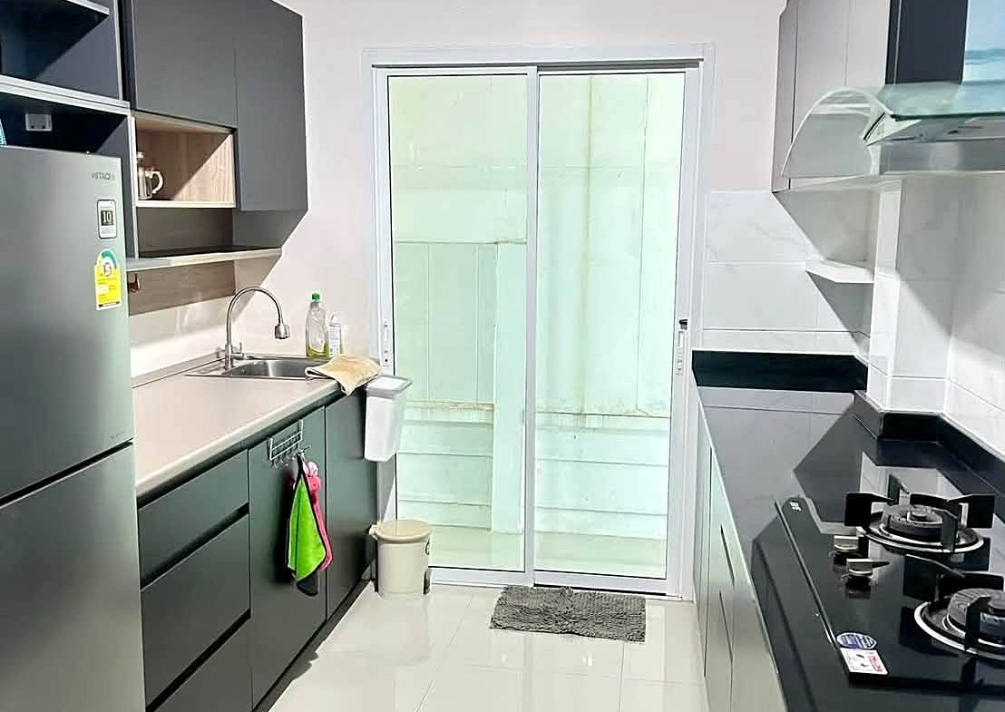 Modern galley kitchen with dark cabinets, stainless fridge, sink on the left, gas cooktop and hood on the right, glass sliding door at the end.