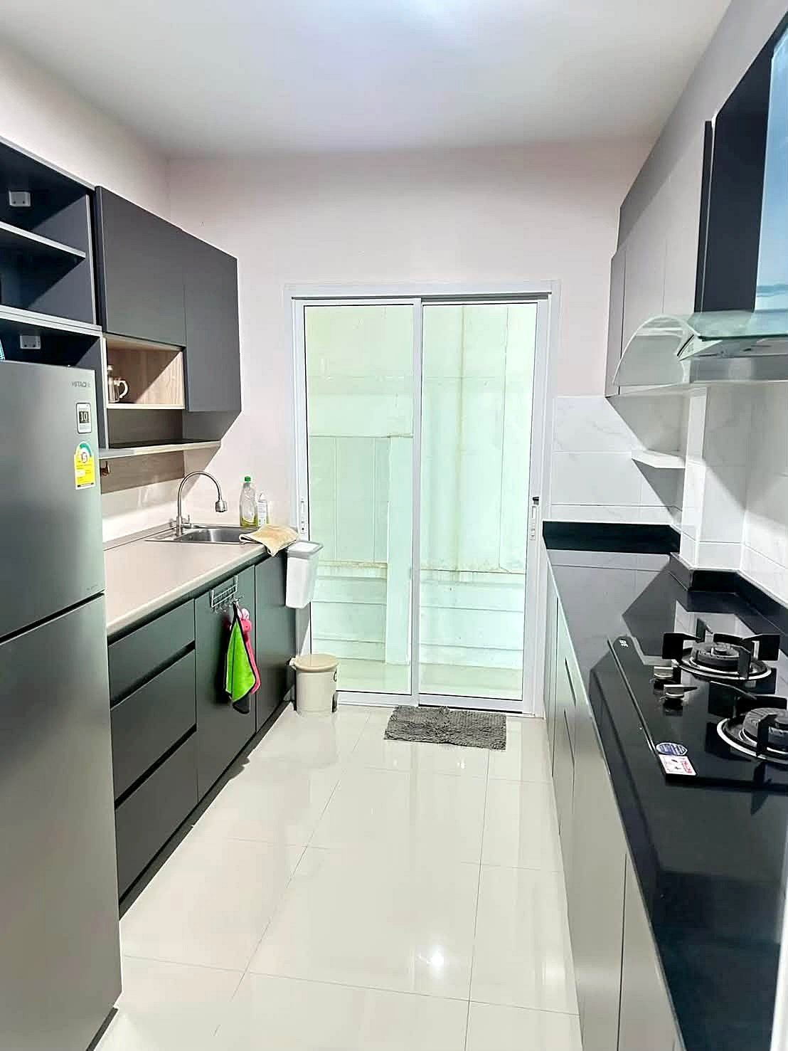 Raviporn_Romsai_Village_Dlh580_11 Modern galley kitchen with dark cabinets, stainless fridge, sink on the left, gas cooktop and hood on the right, glass sliding door at the end.