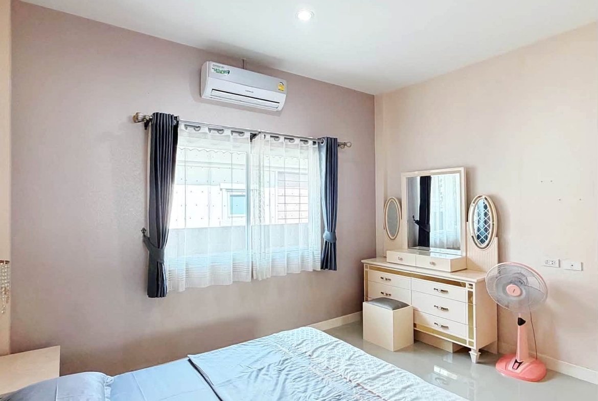 Bedroom with pale walls, window dressed in sheer white and dark curtains, an air conditioner above the window, and a white dresser with two oval mirrors; a pink standing fan sits nearby with a small stool in front of the dresser.