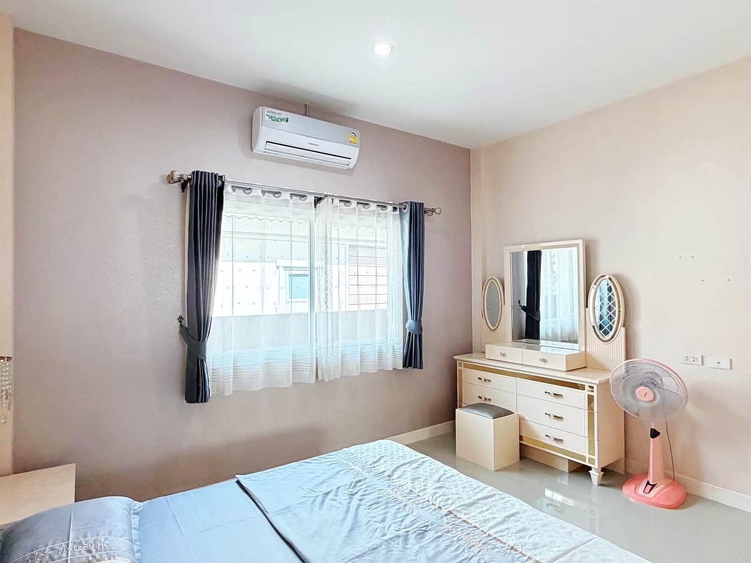 Raviporn_Romsai_Village_Dlh580_15 Bedroom with pale walls, window dressed in sheer white and dark curtains, an air conditioner above the window, and a white dresser with two oval mirrors; a pink standing fan sits nearby with a small stool in front of the dresser.