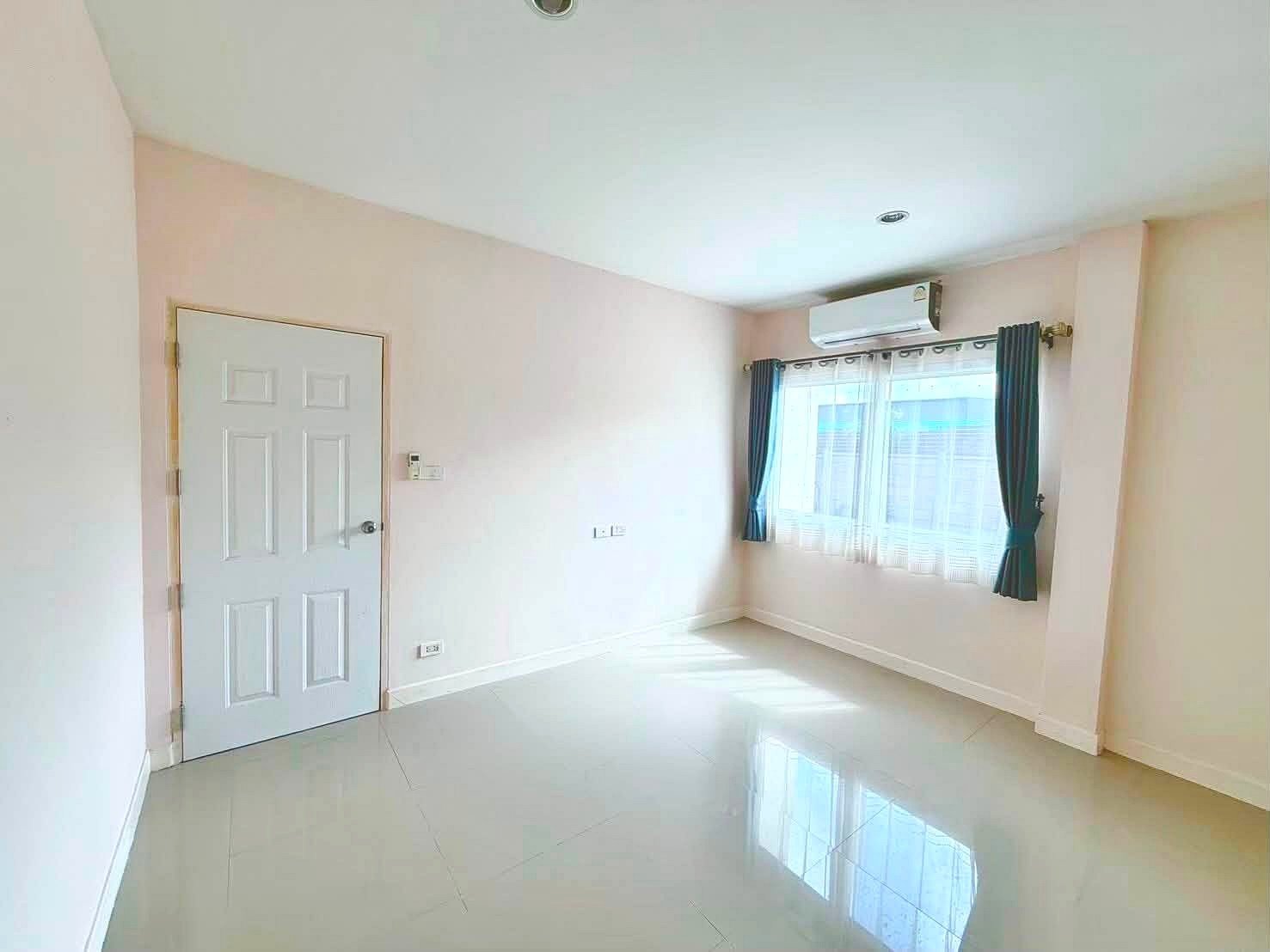 Raviporn_Romsai_Village_Dlh580_18 Empty bright room with pale pink walls, white door, large window with blue curtains, and a wall-mounted air conditioner above the window.