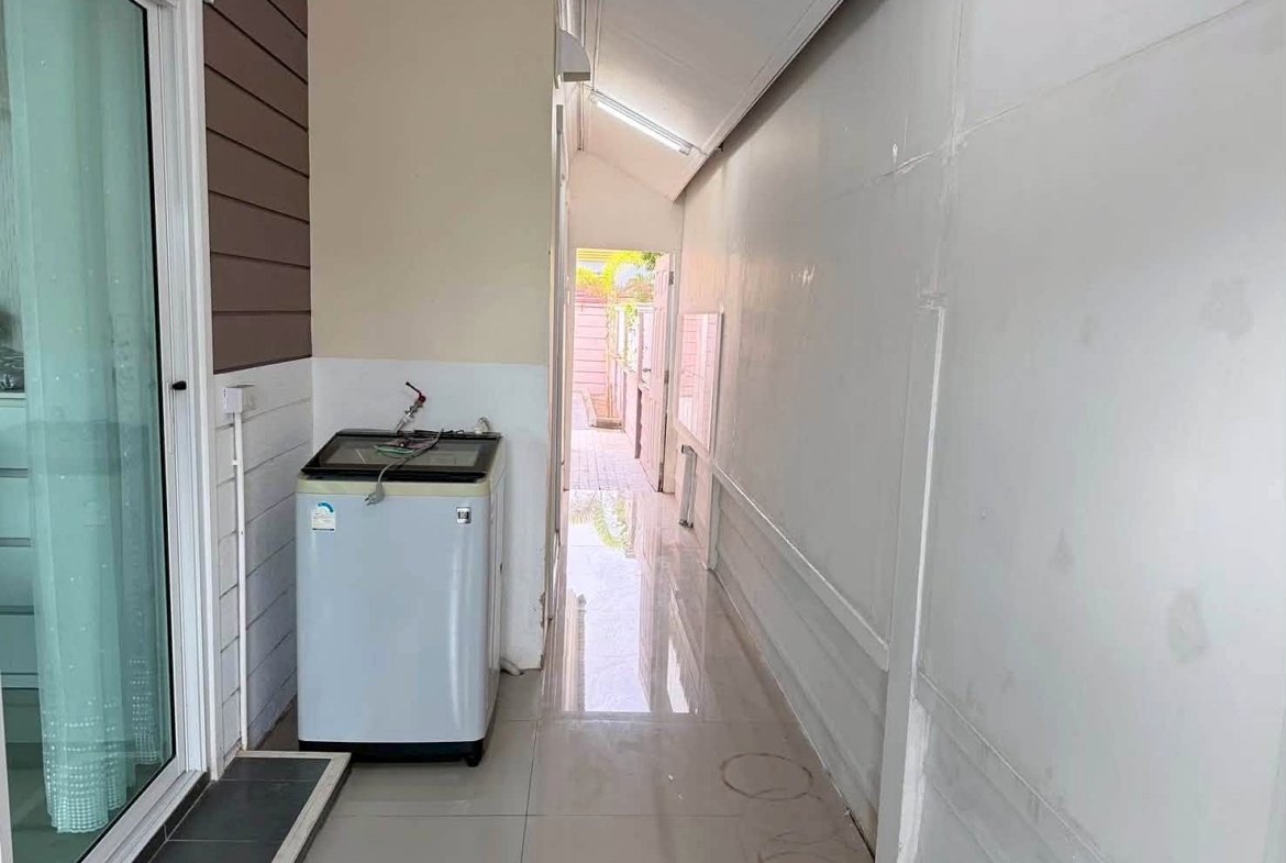 Narrow outdoor laundry area beside a sliding glass door; washing machine against a beige wall with wood siding on the left and a white plastic-covered wall on the right.