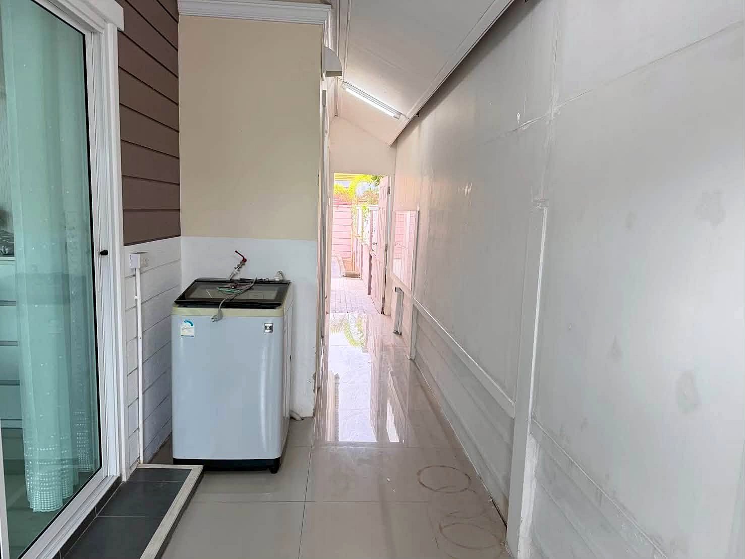 Raviporn_Romsai_Village_Dlh580_22 Narrow outdoor laundry area beside a sliding glass door; washing machine against a beige wall with wood siding on the left and a white plastic-covered wall on the right.
