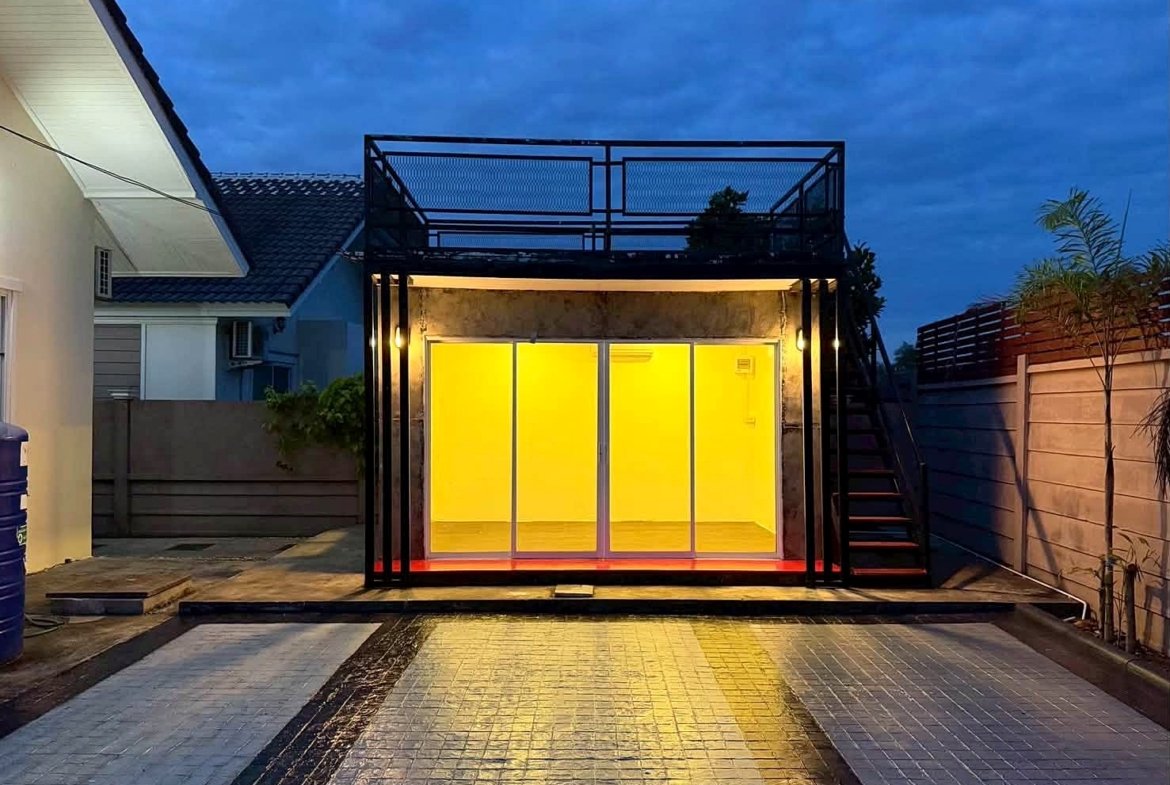 Modern outdoor glass room with bright yellow interior lighting, encased in a black metal frame and a rooftop deck above.