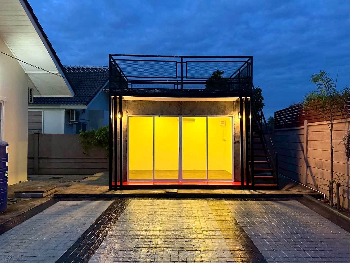 Raviporn_Romsai_Village_Dlh580_26 Modern outdoor glass room with bright yellow interior lighting, encased in a black metal frame and a rooftop deck above.