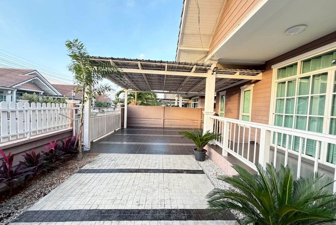 Front walkway of a modern house with a tiled path, white fence, and a covered carport, with potted plants along the sides.