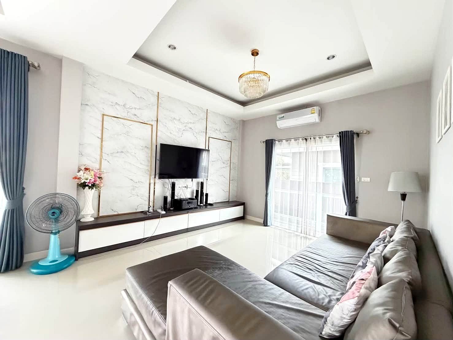 Raviporn_Romsai_Village_Dlh580_3 Bright modern living room with a gray sectional sofa, marble-panel TV wall, and a wall-mounted TV above a white storage console.