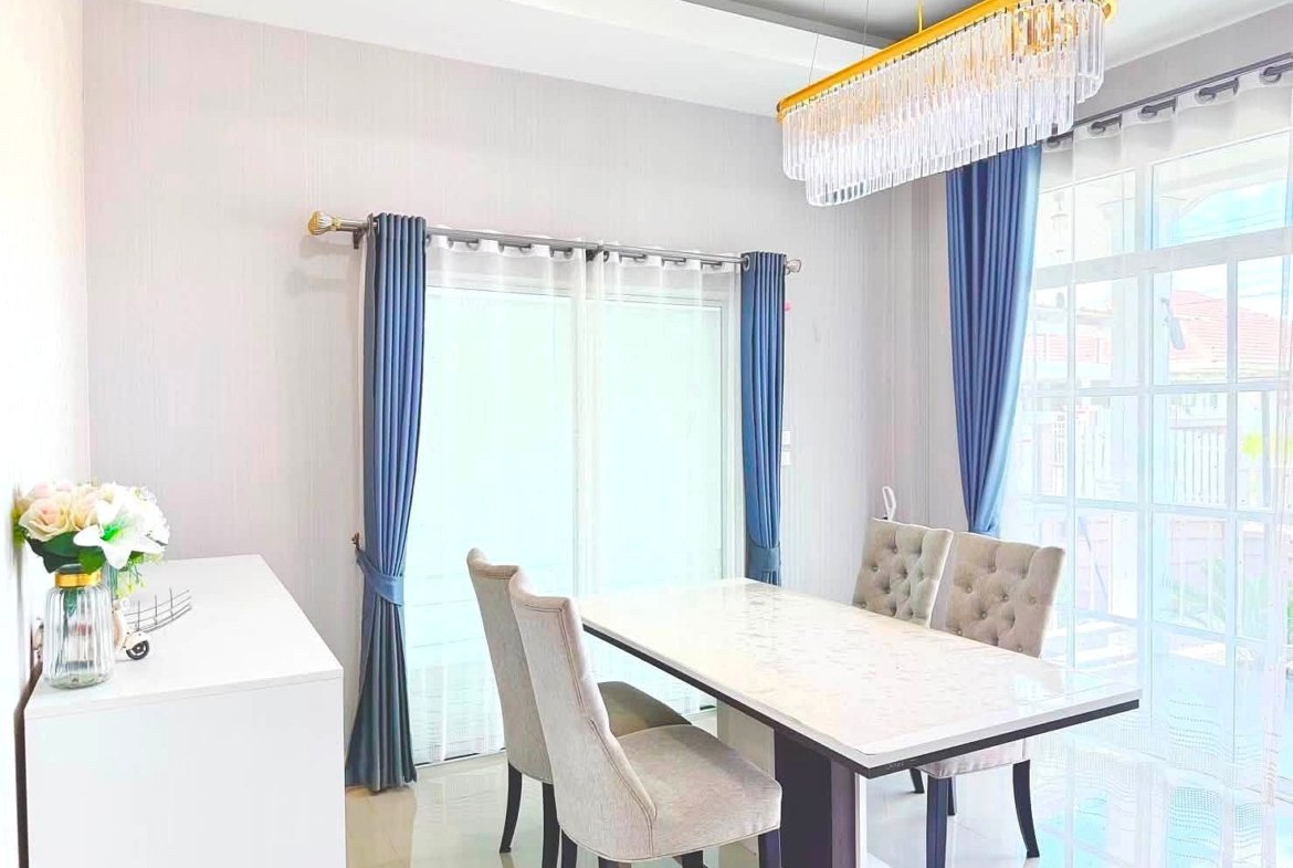 Bright dining room with a white marble table, tufted beige chairs, blue curtains, and a crystal chandelier above.