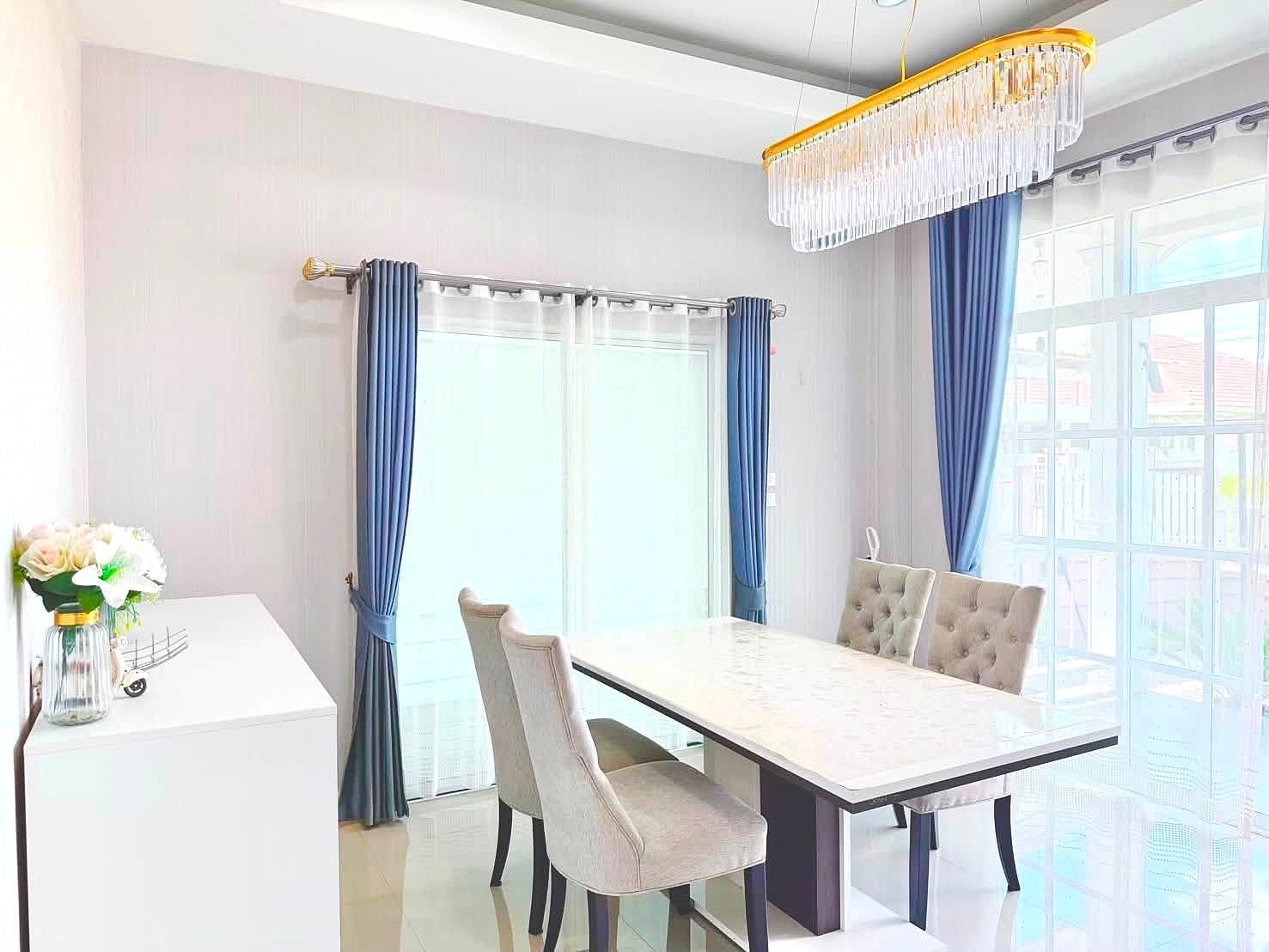 Raviporn_Romsai_Village_Dlh580_4 Bright dining room with a white marble table, tufted beige chairs, blue curtains, and a crystal chandelier above.