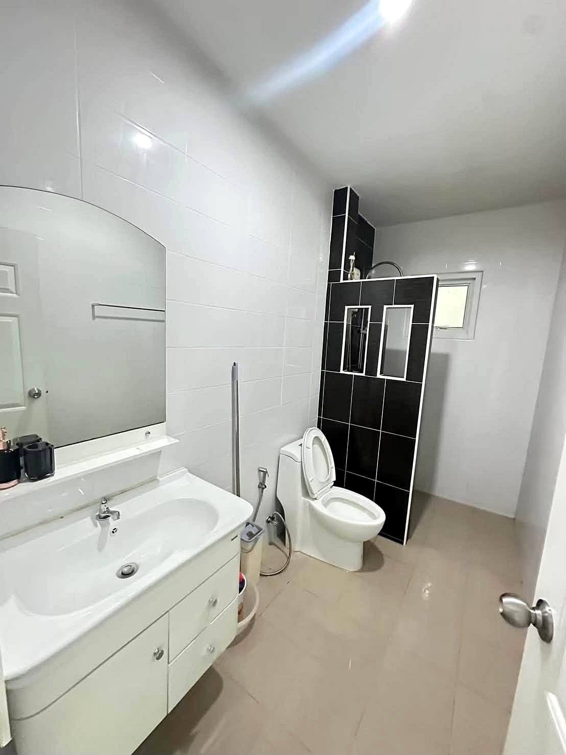 Raviporn_Romsai_Village_Dlh580_8 Modern bathroom with white vanity, oval mirror, and toilet; black tiled shower divider in the corner.