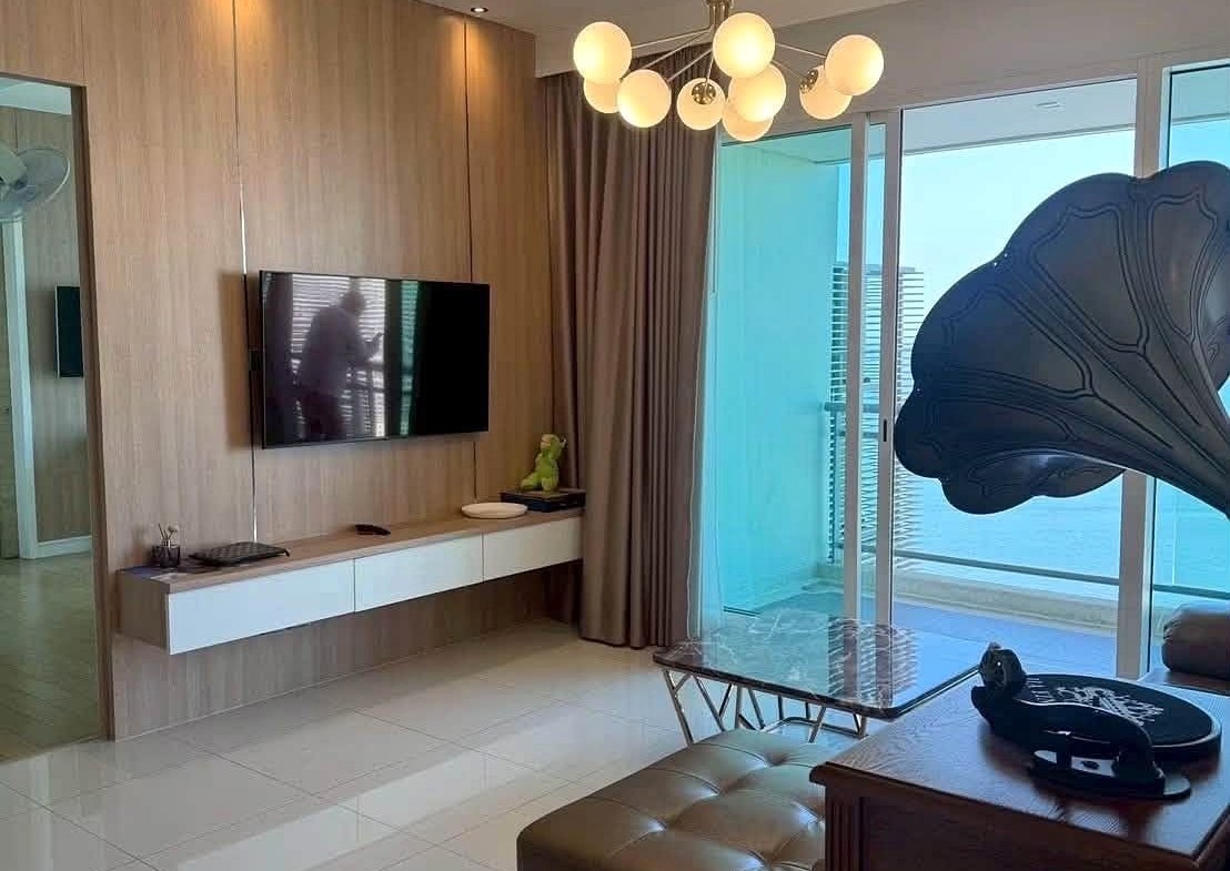 Living room with a wall-mounted TV on a wood-paneled feature wall, white floating console, and a balcony beyond glass doors in view.