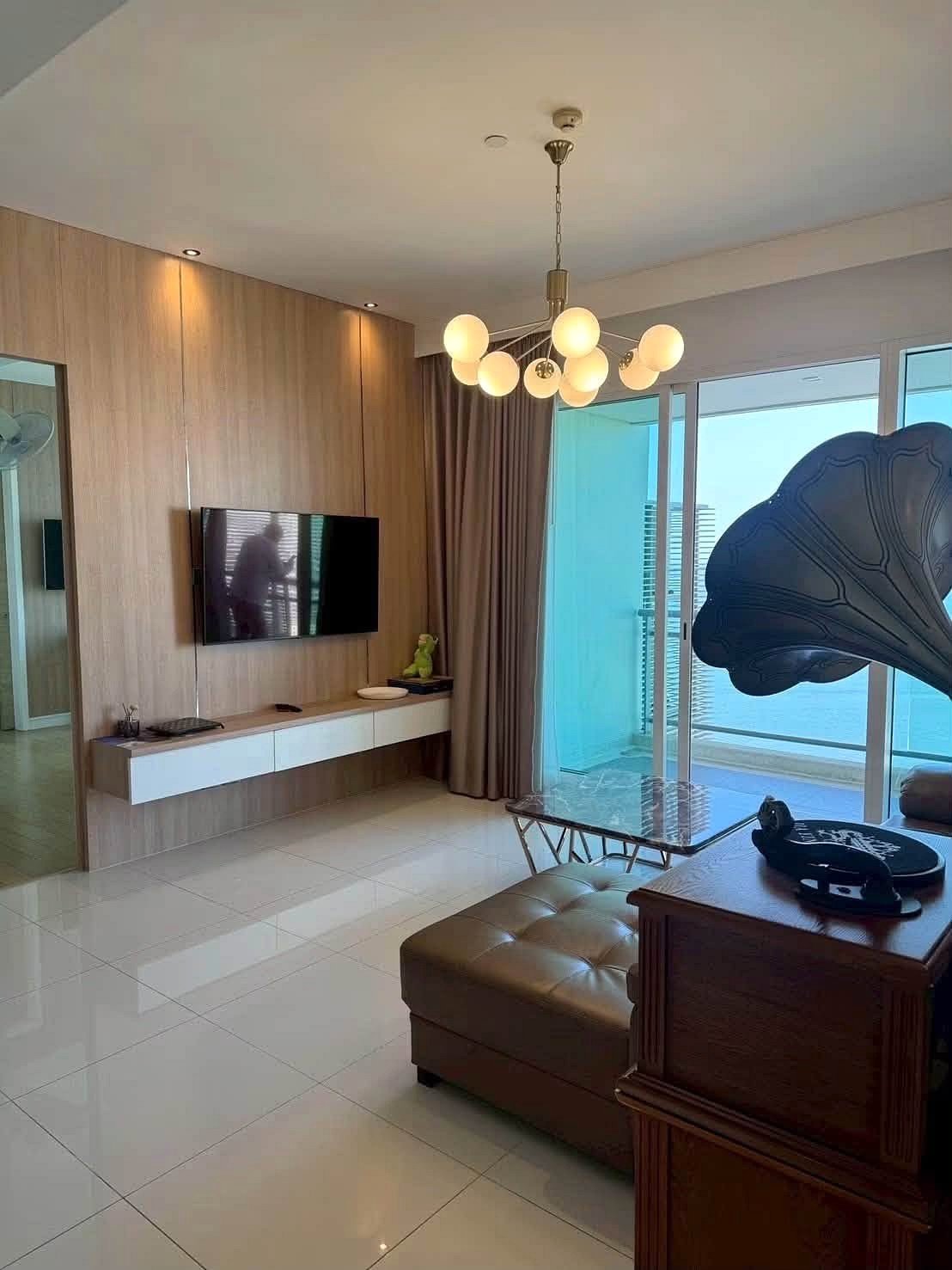 Reflectiom_Jomtien_2Bed_Dlc625 Living room with a wall-mounted TV on a wood-paneled feature wall, white floating console, and a balcony beyond glass doors in view.