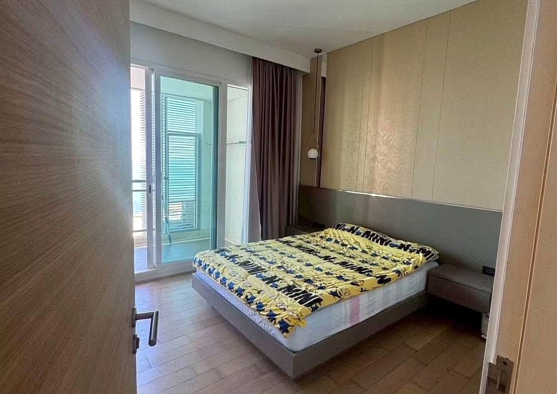 Hotel bedroom with a low platform bed, yellow-blue patterned quilt, and gray bedside tables under a beige paneled wall near a balcony door.