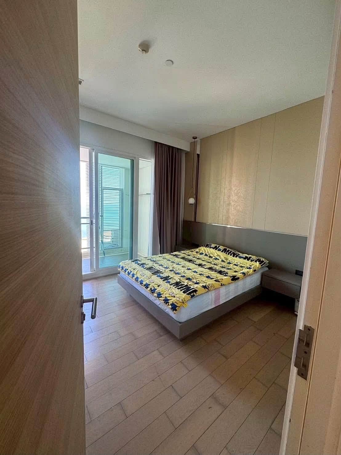 Reflectiom_Jomtien_2Bed_Dlc625_12 Hotel bedroom with a low platform bed, yellow-blue patterned quilt, and gray bedside tables under a beige paneled wall near a balcony door.