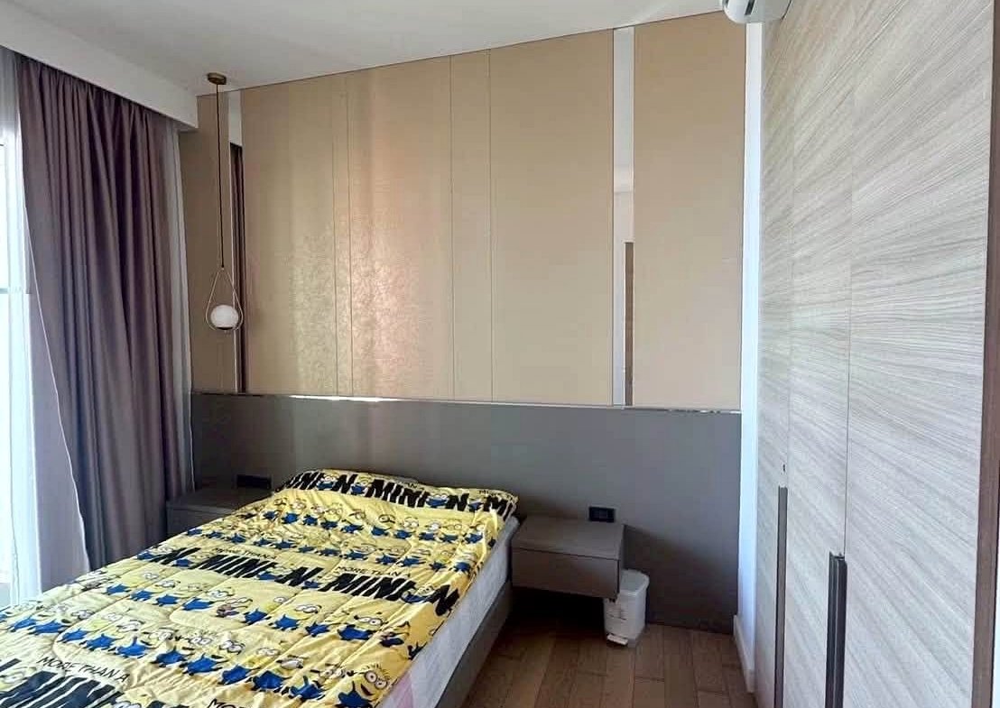 Small modern bedroom with a single bed, gray headboard, and yellow Minions blanket, set against built-in beige wardrobes and a pendant light above. Magnified windows with purple curtains on the left.