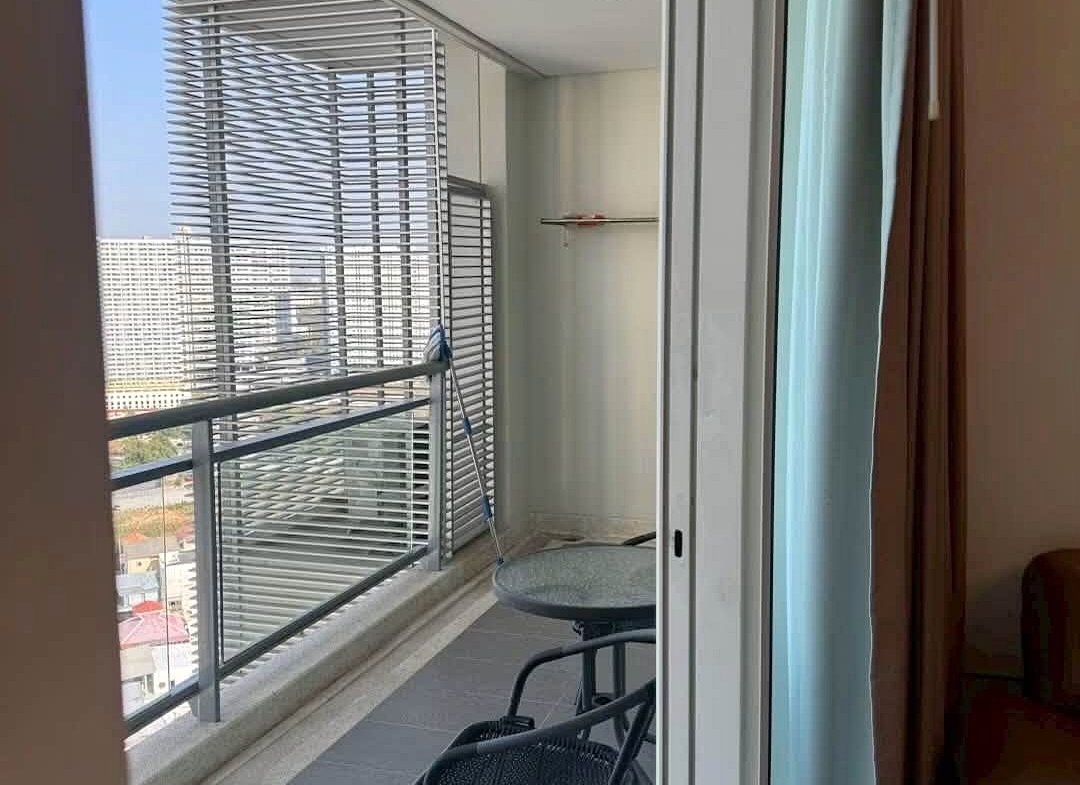 Balcony with metal railing and vertical blinds, a small round glass table and two black chairs, looking out over city buildings from a high floor.