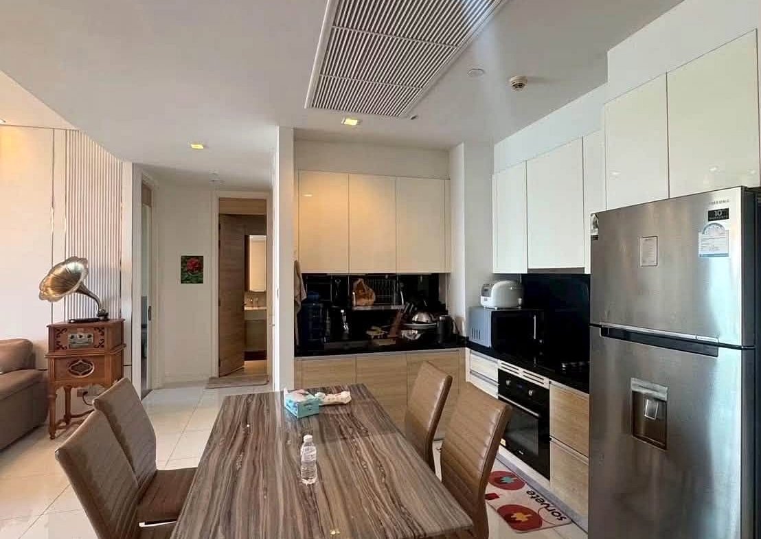 Open-plan kitchen and dining area with a wood dining table and six brown upholstered chairs, stainless steel fridge, white cabinets, and a countertop edge in the background.