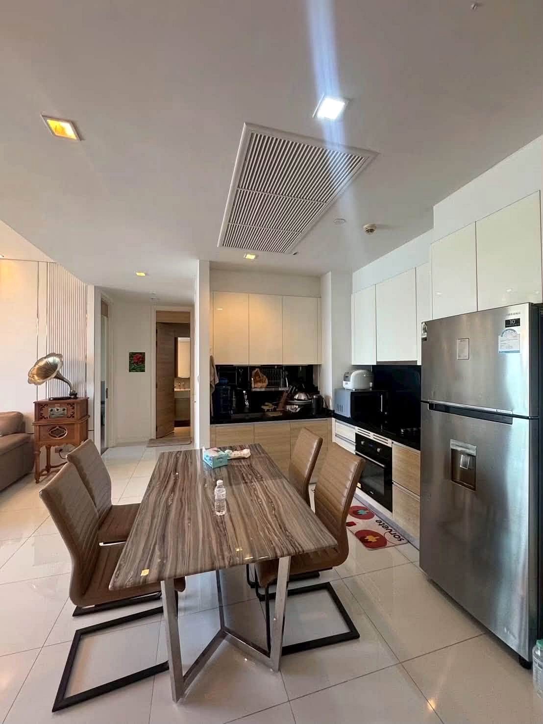 Reflectiom_Jomtien_2Bed_Dlc625_2 Open-plan kitchen and dining area with a wood dining table and six brown upholstered chairs, stainless steel fridge, white cabinets, and a countertop edge in the background.