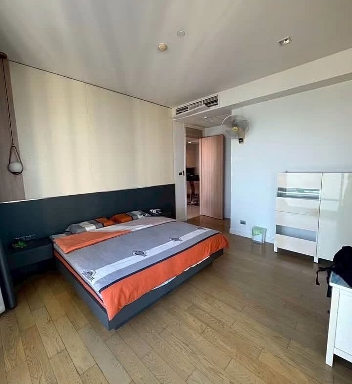 Hotel room with a large bed, orange-gray bedding, and a white dresser against a light wall; wooden floor and open doorway revealing another room.