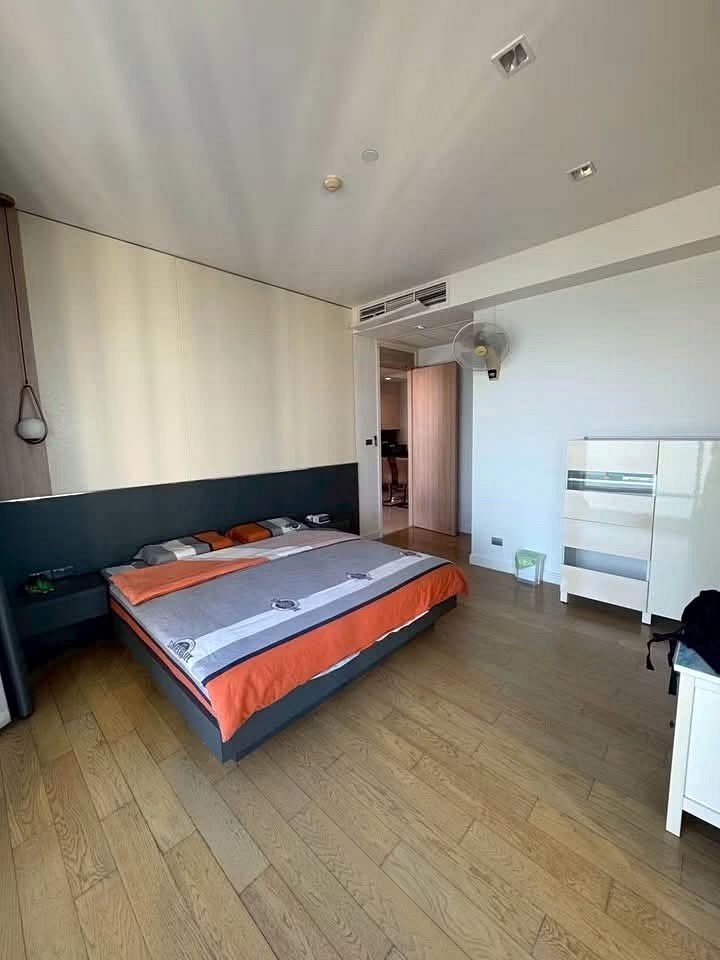 Reflectiom_Jomtien_2Bed_Dlc625_3 Hotel room with a large bed, orange-gray bedding, and a white dresser against a light wall; wooden floor and open doorway revealing another room.