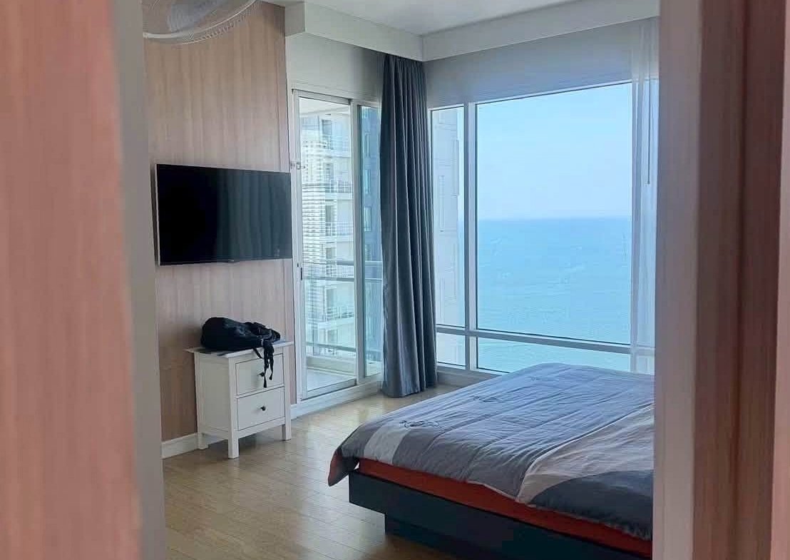 Bedroom with a sea view: large windows, bed, blue-gray bedding, and a wall-mounted TV on a wooden panel wall.
