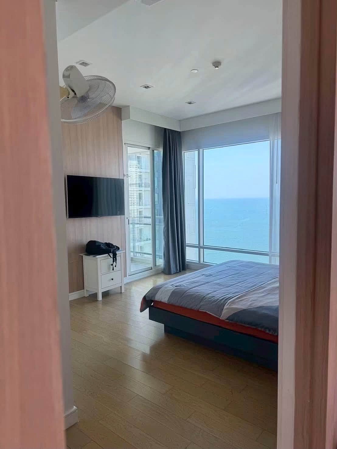 Reflectiom_Jomtien_2Bed_Dlc625_4 Bedroom with a sea view: large windows, bed, blue-gray bedding, and a wall-mounted TV on a wooden panel wall.