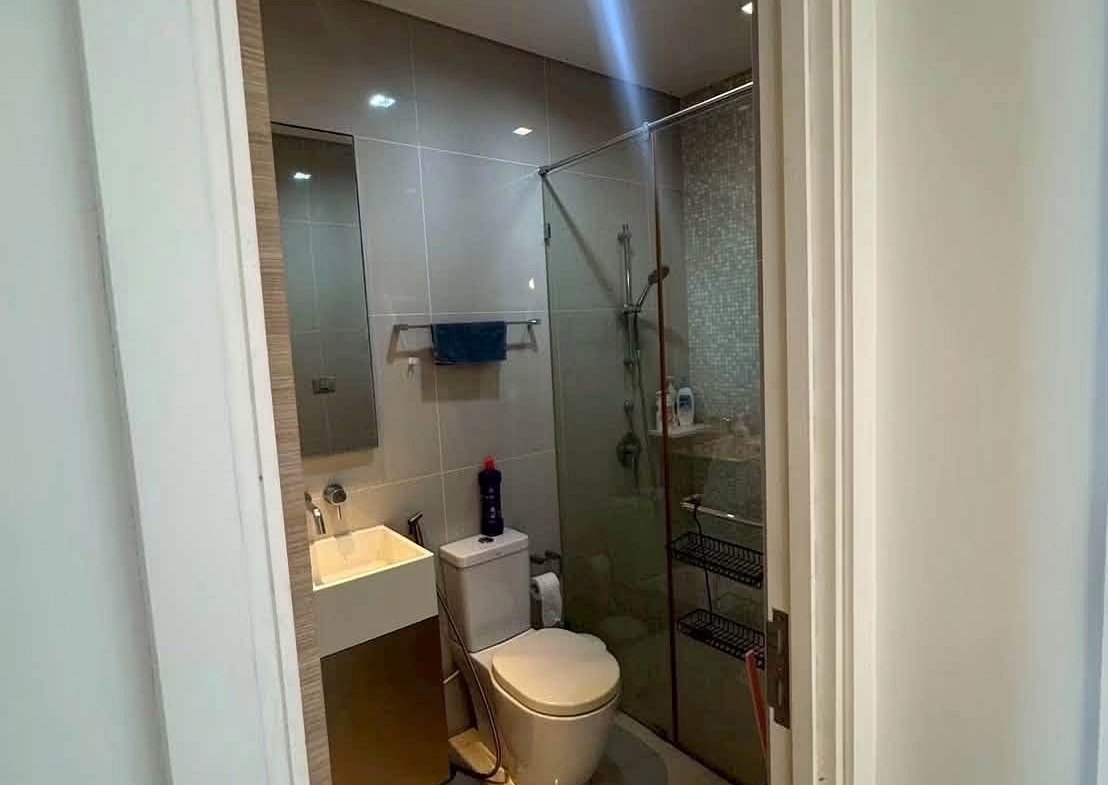 Small modern bathroom featuring a glass shower enclosure, toilet, and vanity with white sink; blue towel on a wall rack and a bottle on the toilet tank visible through the doorway.