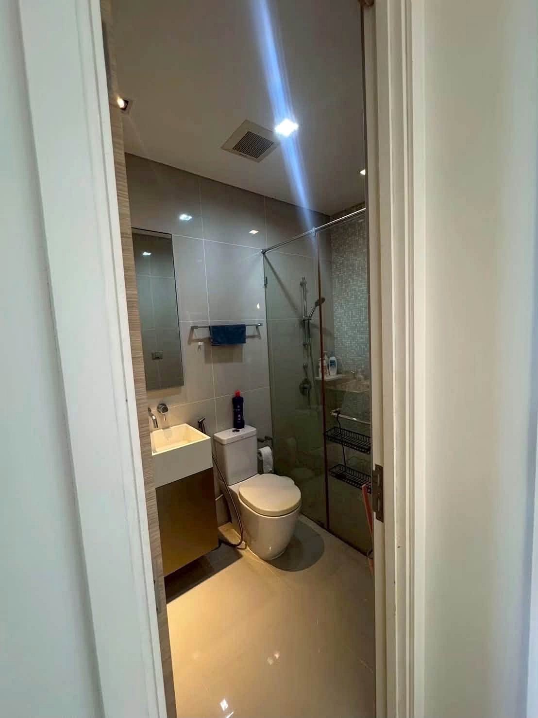 Reflectiom_Jomtien_2Bed_Dlc625_5 Small modern bathroom featuring a glass shower enclosure, toilet, and vanity with white sink; blue towel on a wall rack and a bottle on the toilet tank visible through the doorway.