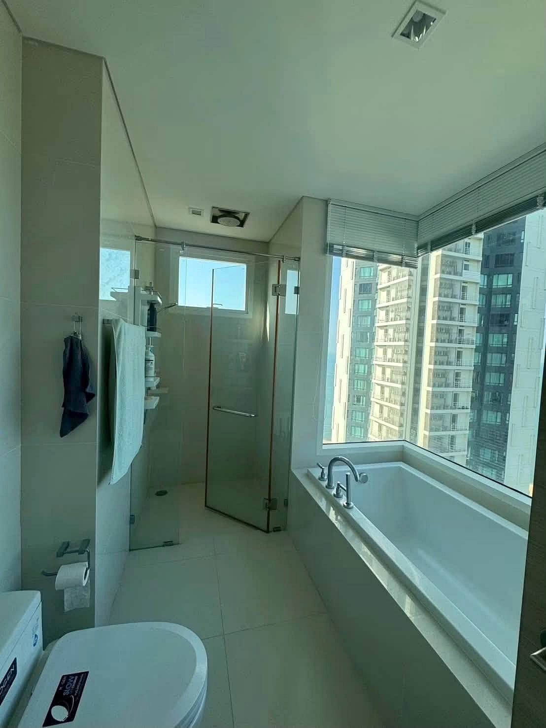 Reflectiom_Jomtien_2Bed_Dlc625_6 Bright, modern bathroom with a glass-enclosed shower and a white tub by a large window overlooking city high-rises.