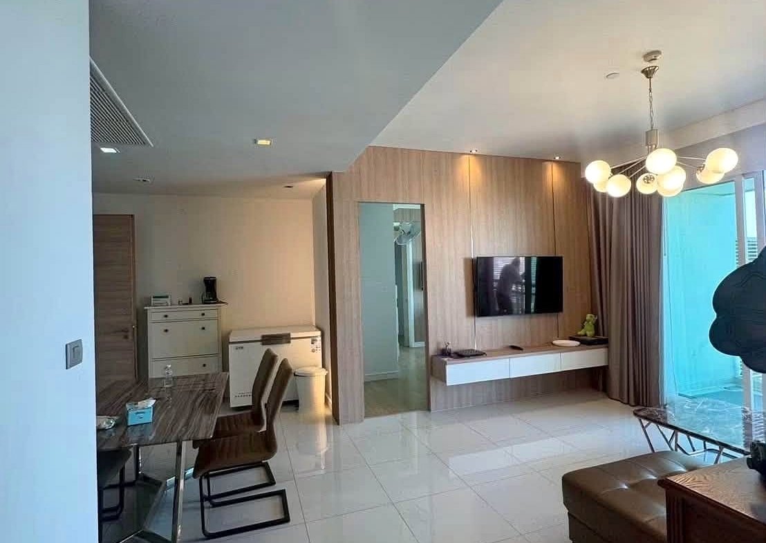 Open-plan living area with a wall-mounted TV on a wood-paneled wall and a glass dining table with brown chairs nearby.