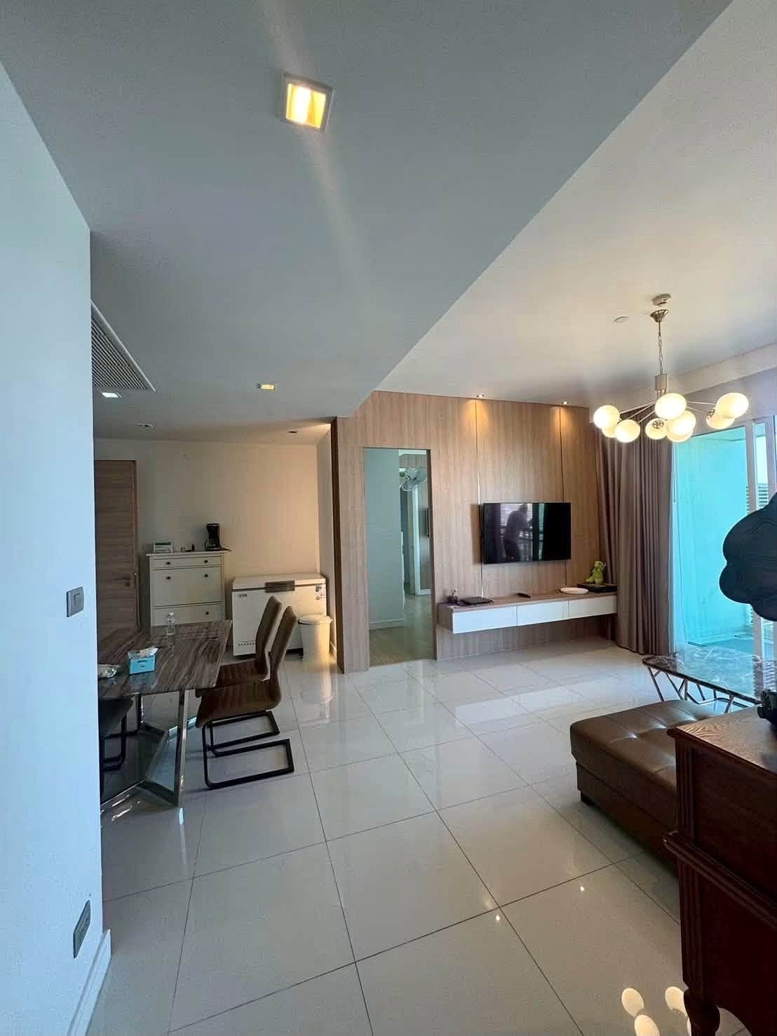 Reflectiom_Jomtien_2Bed_Dlc625_9 Open-plan living area with a wall-mounted TV on a wood-paneled wall and a glass dining table with brown chairs nearby.